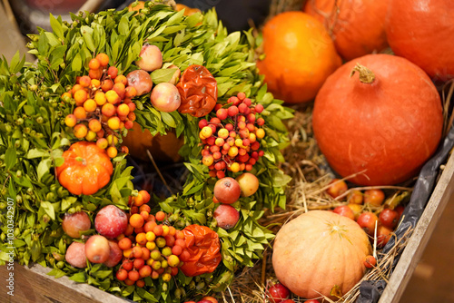 Eine lebendige Herbstgestaltung zeigt einen Kranz aus grünen Blättern, kleinen roten und orangefarbenen Beeren, Äpfeln und umgeben von Kürbissen auf einem Strohbett