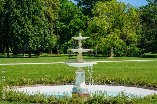 A classic white fountain gracefully flows in the center of a lush, green park surrounded by tall trees
