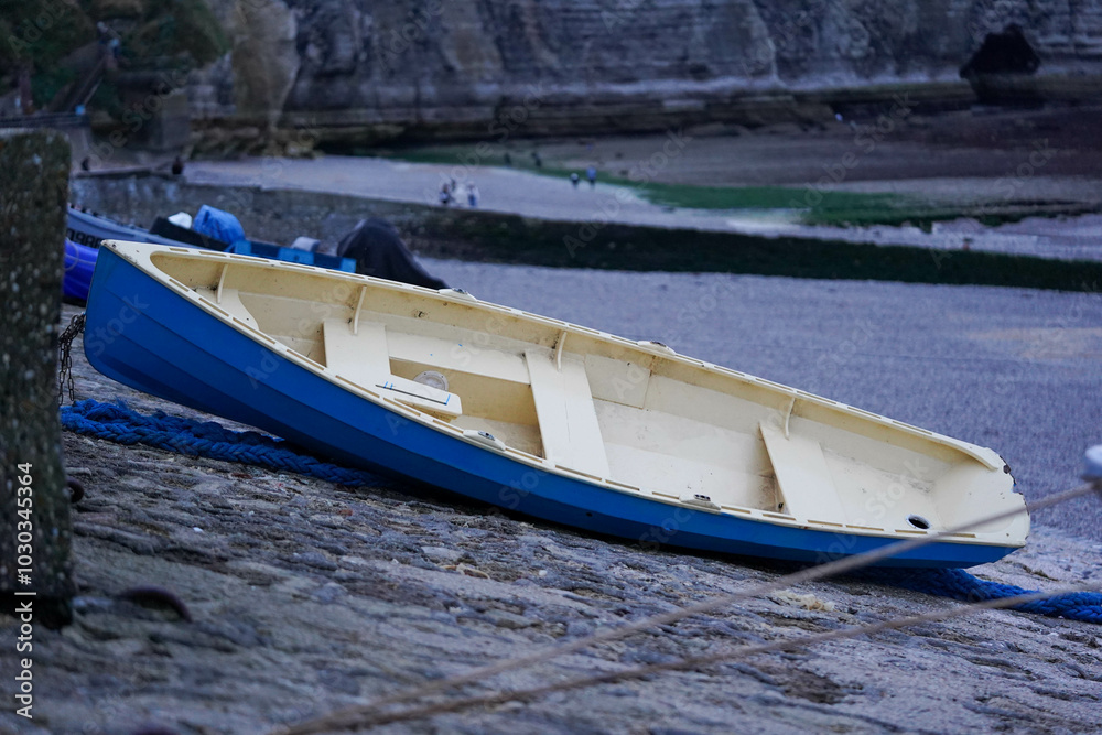 A small blue and beige boat is resting upside down on a stone surface near the shore, with a calm beach and distant cliffs in the background A small blue and beige boat is resting upside down on a stone surface near the shore, with a calm beach and distant cliffs in the background