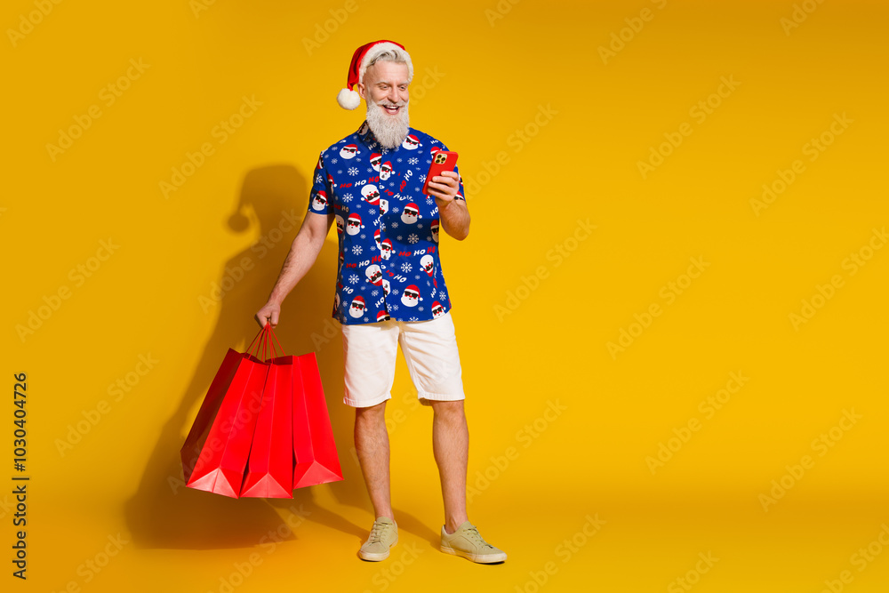 Full size photo of stylish modern santa smart phone shop bags empty space xmas winter print shirt isolated on yellow color background Full size photo of stylish modern santa smart phone shop bags empty space xmas winter print shirt isolated on yellow color background
