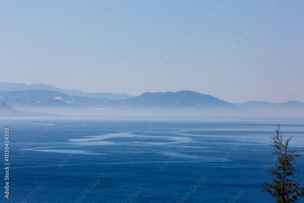 Early mornings view to the sea  and blue mountains on horizon. Slight haze above the surface of the sea and path of wind on water.  Top of pine in right part of picture