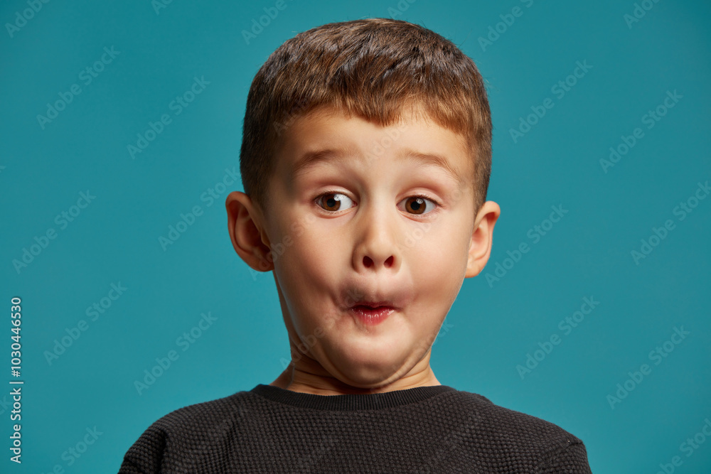 Boy makes comical expression with puffed cheeks, his eyes wide open in surprise against blue studio background. Childhood curiosity. Concept of facial-expressions, positive and negative emotions. Ad Boy makes comical expression with puffed cheeks, his eyes wide open in surprise against blue studio background. Childhood curiosity. Concept of facial-expressions, positive and negative emotions. Ad