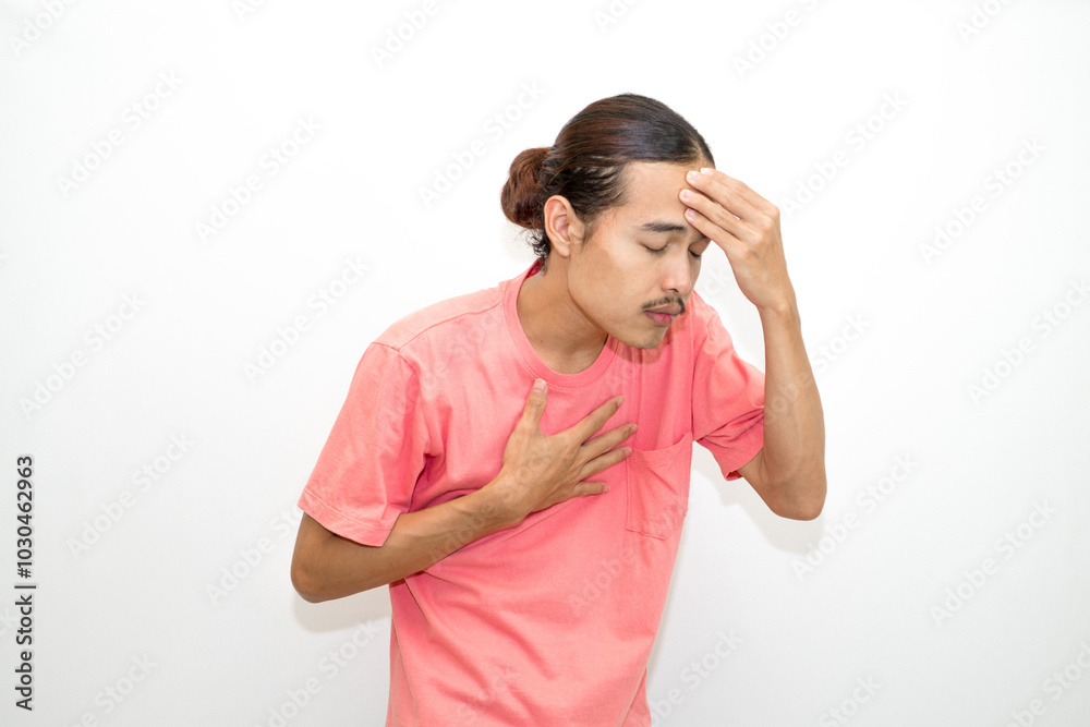 An Indonesian, Asian man wearing a pink tshirt having a headache, chest pain and stomach ache with hands holding head, chest and stomach. isolated in white background An Indonesian, Asian man wearing a pink tshirt having a headache, chest pain and stomach ache with hands holding head, chest and stomach. isolated in white background