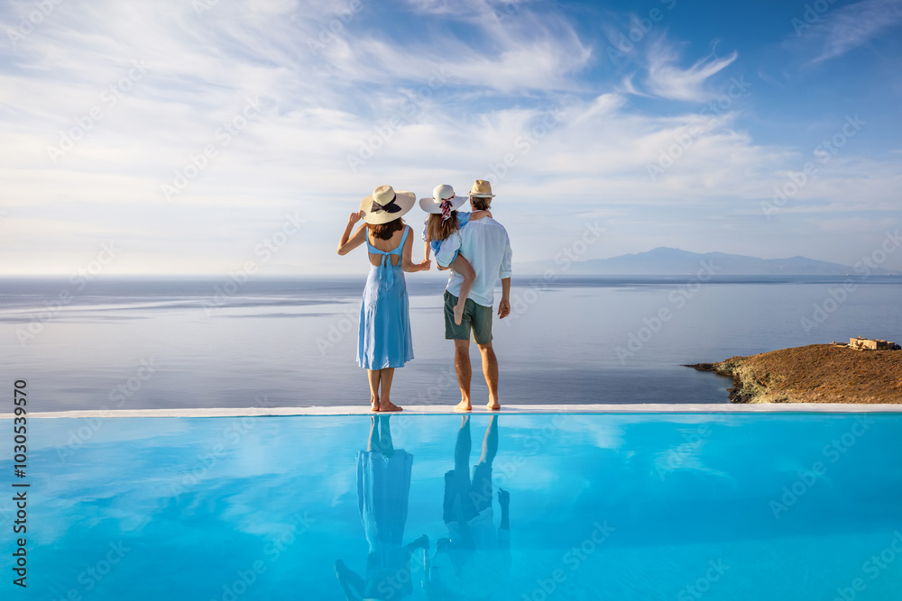 A family on summer holiday time stands at the edge of an infinity swimming pool and enjoys the view over the ocean