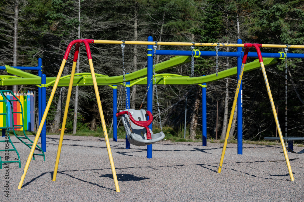 Fototapeta premium A children's playground with a colorful metal swing set, a children's swing chair and a monkey climber. The background is a wooden fence and a wooded area. A climbing structure made of green metal.