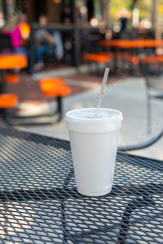 A large or tall white nonrecyclable to-go drink cup with a plastic lid and straw on a black metal patio table. The polystyrene or Styrofoam insulated drinking cup is filled with a cold soda pop drink photo