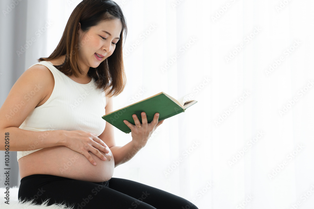 Smiling pregnant Asian young woman sitting and reading book at home while touching her belly with love and care. Pregnancy lifestyle. Copy space Smiling pregnant Asian young woman sitting and reading book at home while touching her belly with love and care. Pregnancy lifestyle. Copy space