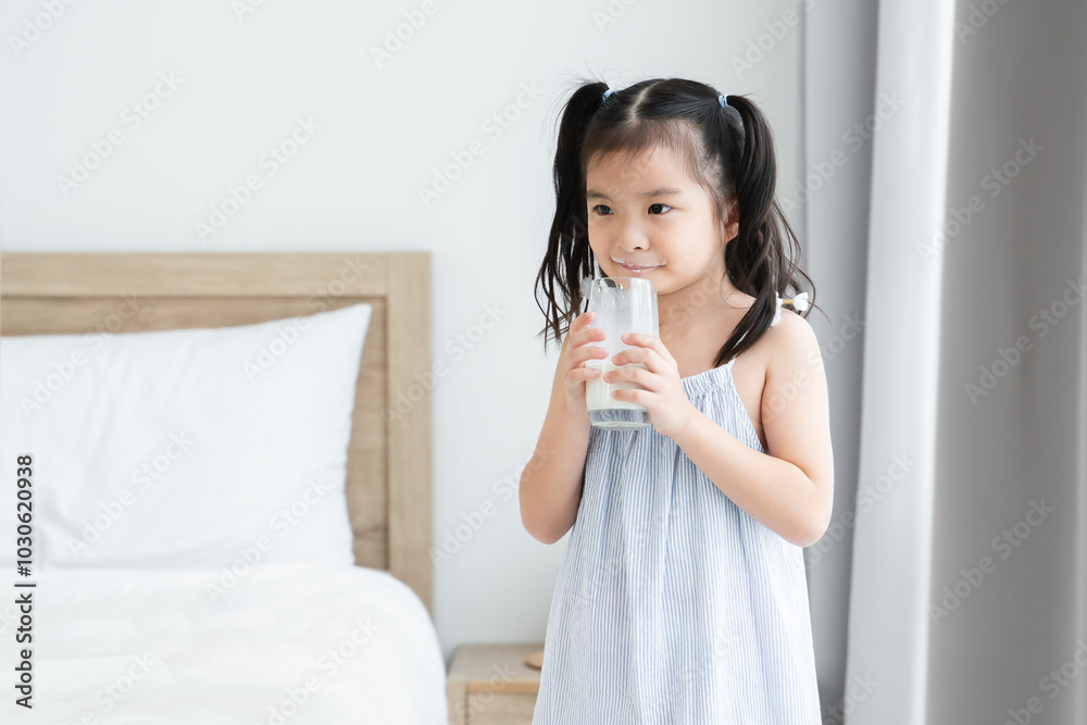 Asian little cute kid with mustache of milk on lips, holding a glass of milk in bedroom at home. Young preschool child girl enjoy drinking milk for good health for breakfast in morning Asian little cute kid with mustache of milk on lips, holding a glass of milk in bedroom at home. Young preschool child girl enjoy drinking milk for good health for breakfast in morning