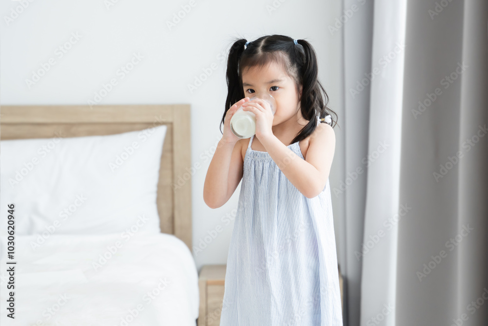 Asian little cute kid drinking while holding a glass of milk, standing in bedroom at home. Young preschool child girl drinking milk for good health for breakfast in morning Asian little cute kid drinking while holding a glass of milk, standing in bedroom at home. Young preschool child girl drinking milk for good health for breakfast in morning