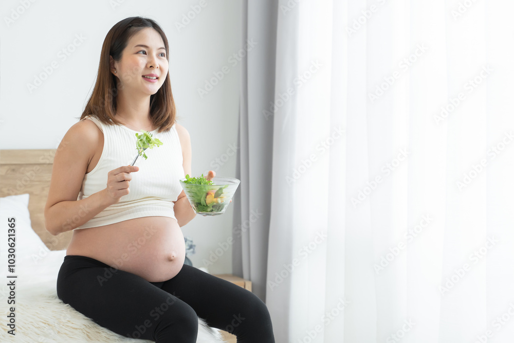 Young pregnant woman is sitting, holding a bowl of salad and fork. She is smiling, looking out of window in bedroom at home. Asian beautiful mother eating fresh vegetable salad for good health Young pregnant woman is sitting, holding a bowl of salad and fork. She is smiling, looking out of window in bedroom at home. Asian beautiful mother eating fresh vegetable salad for good health