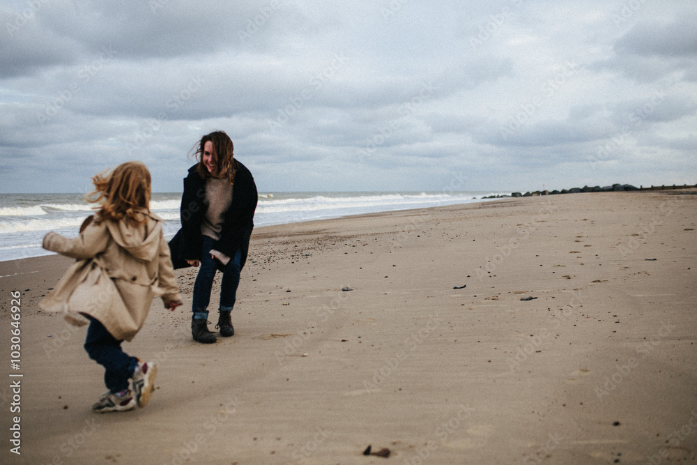 Mother in a warm coat and little daughter in a trench coat run after each other and have fun on the sandy beach with a strong wind in autumn, Norfolk, England