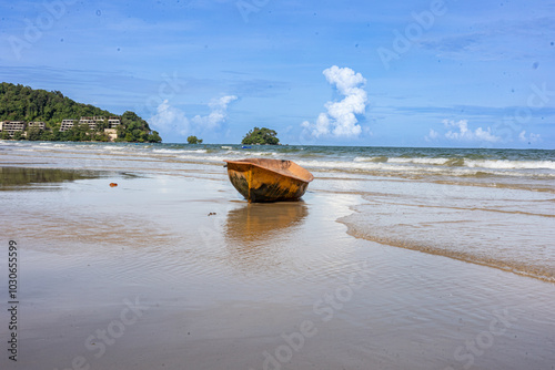 A wooden boat on a sandy beach, partly in the water. The boat, worn and brown, sits at an angle in the turquoise water. The serene scene includes a wide sandy beach, trees, and a cloudy sky. photo