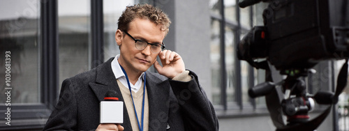 A news anchor in glasses and a coat attentively speaks into a microphone outside a building.