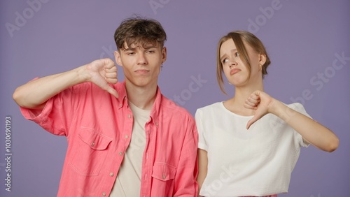 Positive couple of young boyfriend and girlfriend in pink white clothes isolated on purple background, showing thumbs down gesture and sad faces at camera.