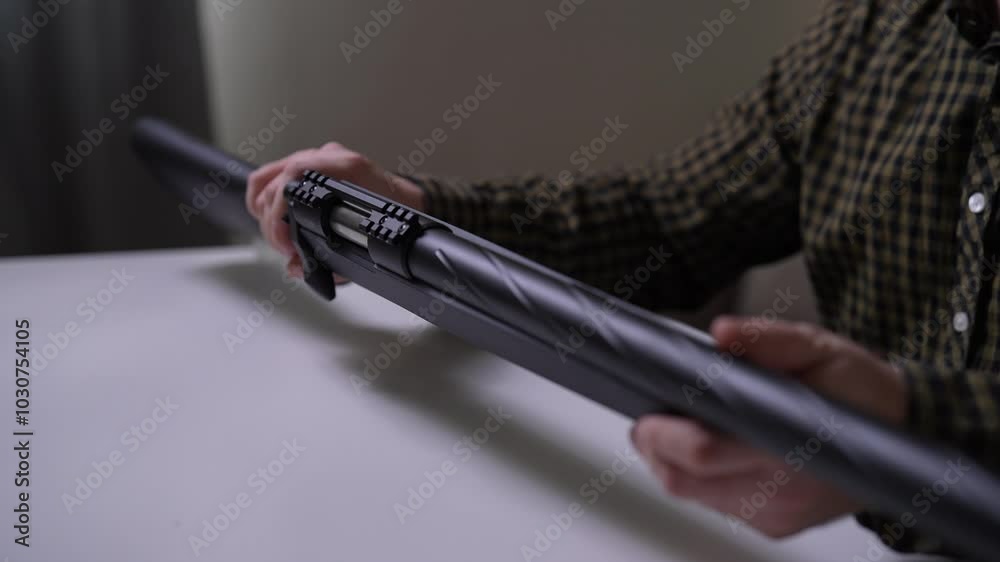 Cropped shot of unrecognizable man in plaid shirt sitting at table, carefully examining black pneumatic sniper rifle, holding gun in various positions, paying attention to features, slow motion. Cropped shot of unrecognizable man in plaid shirt sitting at table, carefully examining black pneumatic sniper rifle, holding gun in various positions, paying attention to features, slow motion.