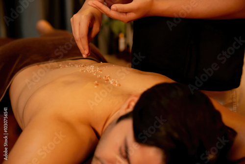 Man customer having exfoliation treatment in luxury spa salon with warmth candle light ambient. Salt scrub beauty treatment in Health spa body scrub. Quiescent