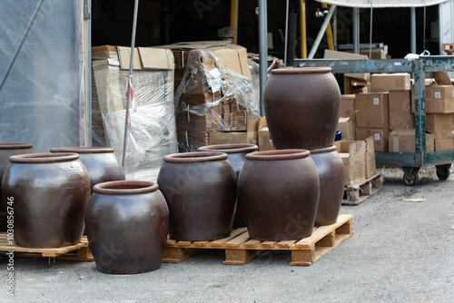 Several ceramic pots are arranged on wooden pallets in an outdoor of a storage facility. Pots vary in size and are crafted from dark clay. Cardboard boxes are visible in the background. Ceramic pots photo