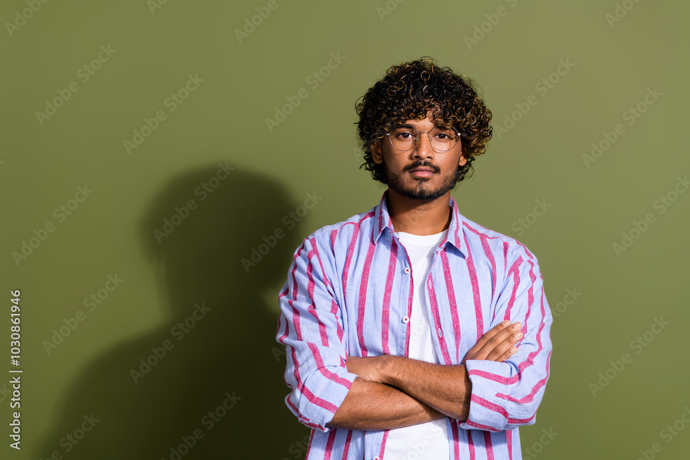Photo of serious confident guy with wavy hair dressed striped shirt in glasses holding arms crossed isolated on khaki color background Photo of serious confident guy with wavy hair dressed striped shirt in glasses holding arms crossed isolated on khaki color background