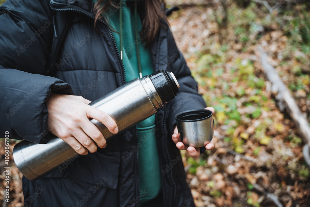 Naklejka premium A joyful woman is seen holding a thermos and a cup, relishing her time outdoors amidst a backdrop of vibrant nature, fully appreciating the stunning beauty that surrounds her