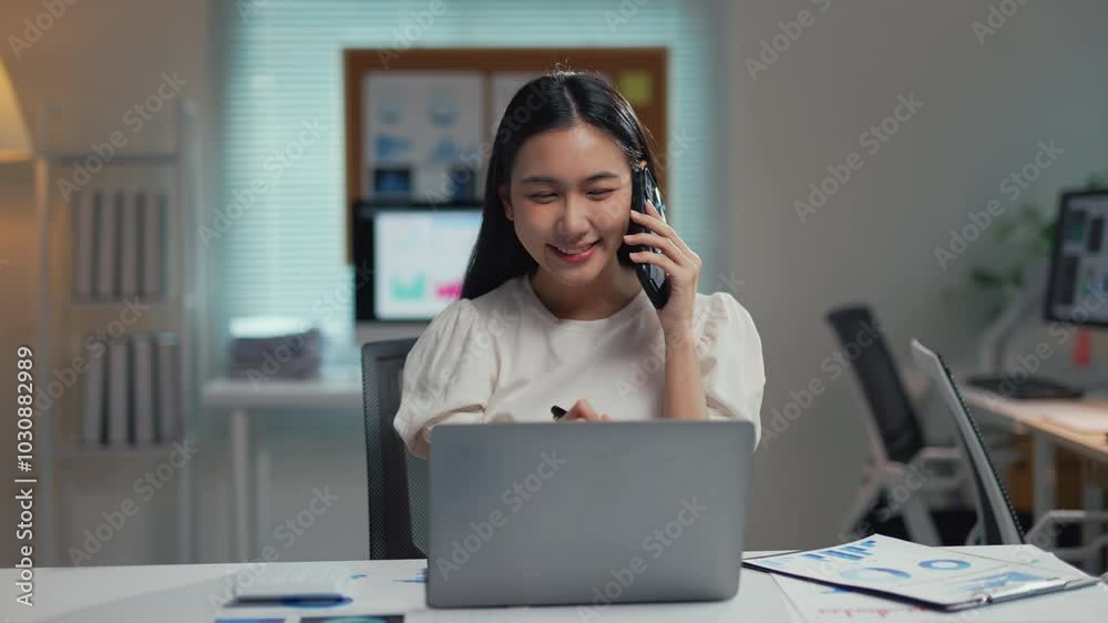 Asian businesswoman is smiling while talking on her phone and working on her laptop at her desk Asian businesswoman is smiling while talking on her phone and working on her laptop at her desk
