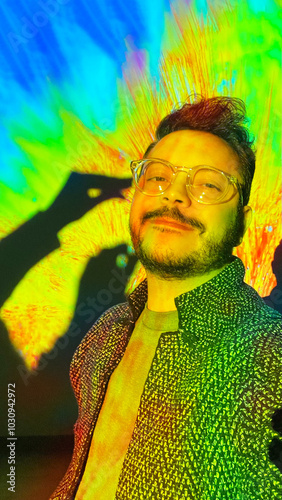 A person wearing glasses smiles for a selfie against a vibrant, colorful background. The playful setting exudes a lively atmosphere, with bright hues enhancing the mood.