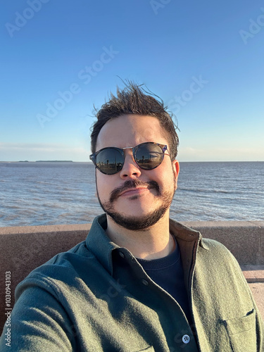 A person wearing sunglasses and a green shirt takes a selfie by the sea on a sunny day. The wind ruffles their hair, and the background features calm water under a clear blue sky.