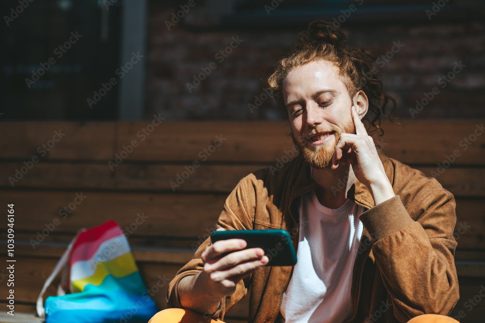 Young man with red hair and a beard happily uses his smartphone on a sunny day outdoors. The image includes an lgbt pride flag symbolizing freedom and support
