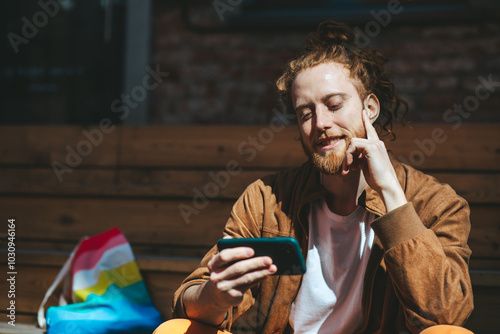 Obraz na płótnie Young man with red hair and a beard happily uses his smartphone on a sunny day outdoors. The image includes an lgbt pride flag symbolizing freedom and support w salonie