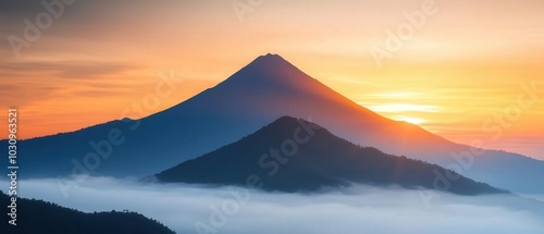 A stunning mountain range at sunset, showcasing a prominent peak with a warm, colorful sky and gentle mist enveloping the lower hills.