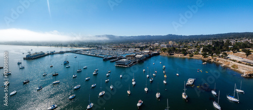 Aerial view of Monterey, California, highlighting a marina with sailboats, piers, and waterfront buildings. Fog covers distant hills under a clear sky.