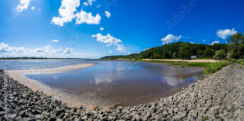 Panoramic view of the Elbe river at Falkensteiner Ufer during low tide with sandy shores, rocky groynes, lush greenery and blue sky, ideal travel destinations and outdoor tourism spot on a summer day.
