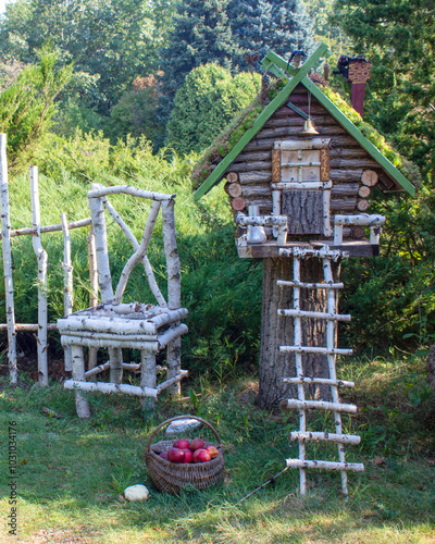 Fairy-tale house made of wooden logs with a ladder made of birch logs. Next to the house is a basket with apples and a chair.