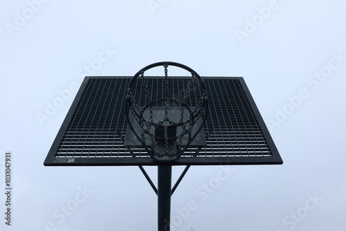 The photo captures a basketball hoop from a low angle, showing a metal backboard and metal net. The sky in the background is gray, adding a stark, industrial feel to the scene. photo