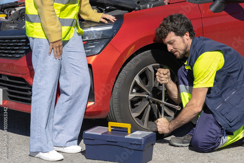 Young professional mechanic changing flat tire broken vehicle on the road with reflective clothing, toolbox and cross wrench, broken engine, roadside assistance, insurance tow truck