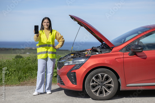 Pretty young woman poses showing phone screen, calling tow truck or insurance mechanic for roadside assistance for car accident, damaged car, broken engine, punctured tire