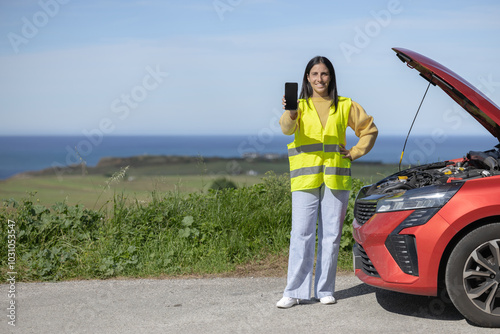 Pretty young woman poses showing phone screen, calling tow truck or insurance mechanic for roadside assistance for car accident, damaged car, broken engine, punctured tire