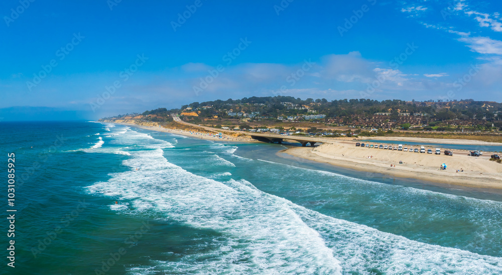 Fototapeta premium Aerial view of San Diego's coastline featuring a sandy beach, a prominent bridge, and a developed area with buildings. Vehicles and people are visible.