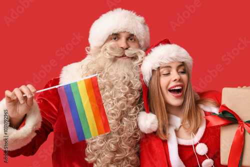 Young couple dressed as Santa Clauses with LGBT flag and Christmas gift on red background