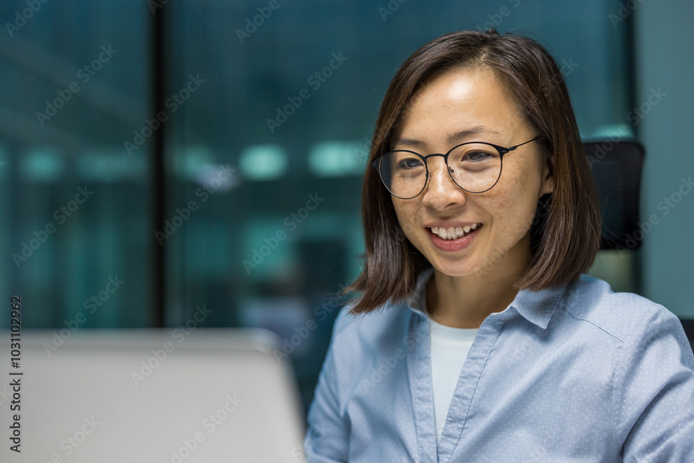 Asian businesswoman working on laptop, smiling confidently in modern office. Image captures essence of digital work, productivity, technology, corporate environment.