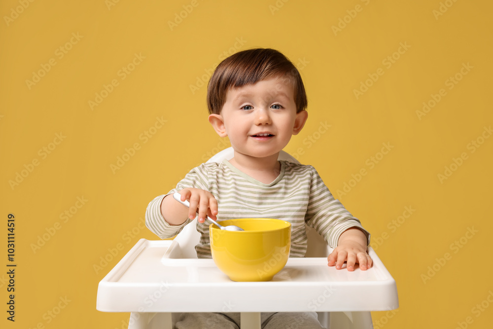 Cute little kid eating healthy baby food from bowl in high chair on yellow background Cute little kid eating healthy baby food from bowl in high chair on yellow background