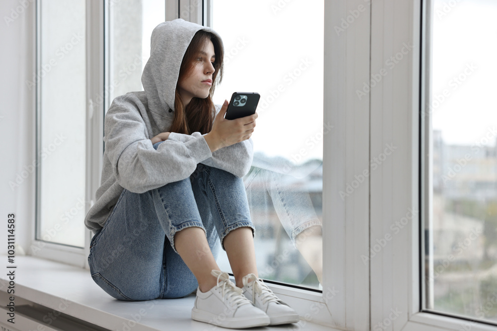 Loneliness concept. Sad teenage girl using smartphone on windowsill at home Loneliness concept. Sad teenage girl using smartphone on windowsill at home