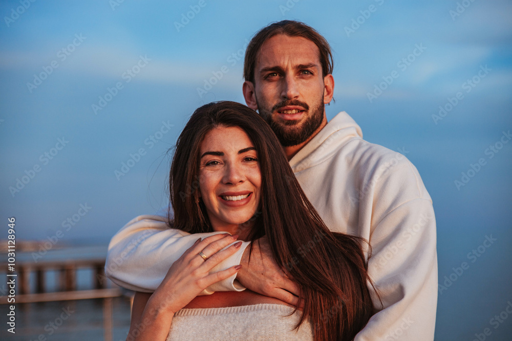 young engaged couple hugging and looking at the camera smiling. They are both dressed in white and are standing on the shore of a beach at sunset. young engaged couple hugging and looking at the camera smiling. They are both dressed in white and are standing on the shore of a beach at sunset.