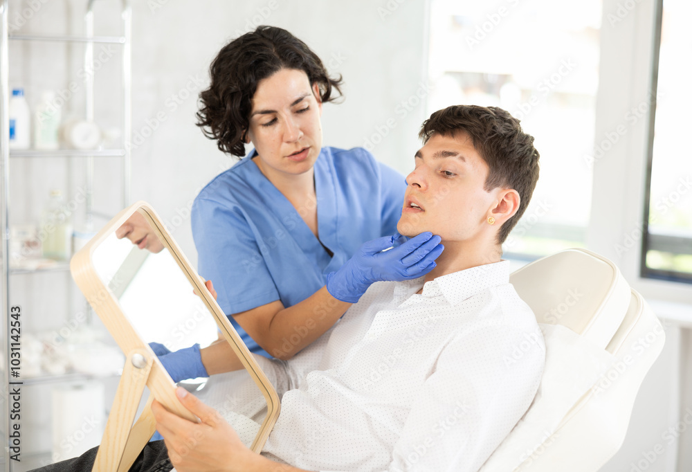 Fototapeta premium Young male patient lying on clinical chair discussing face aesthetic procedures with doctor