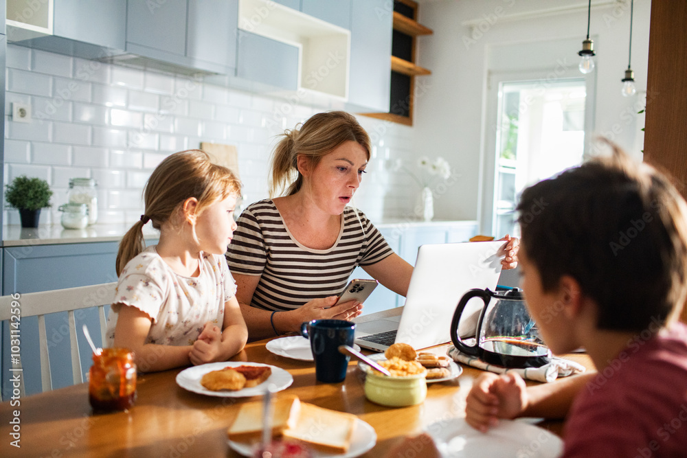 Mother working on laptop while family has breakfast in kitchen Mother working on laptop while family has breakfast in kitchen