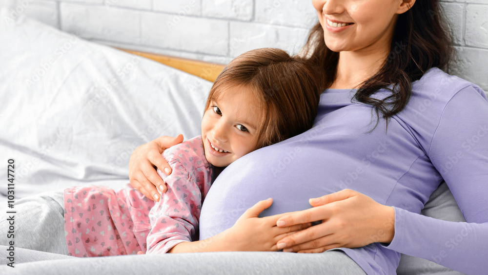 Little daughter and pergnant mother in pajamas bonding on bed, girl hugging mommy tummy, copy space