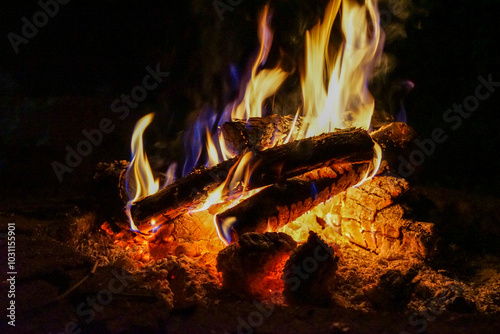 Campfire with yellow and blue flames above glowing bed of orange and white ashes and embers in landscape orientation
