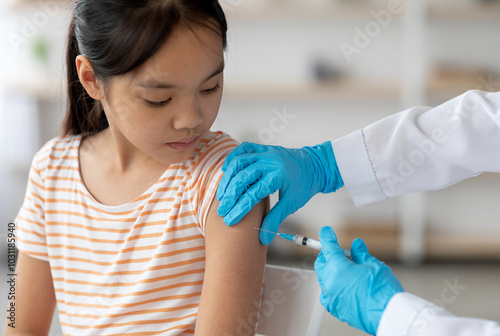 Bild auf Leinwand Unrecognizable doctor hands in protective gloves making vaccination against coronavirus of flu for little asian girl, home interior, closeup shot