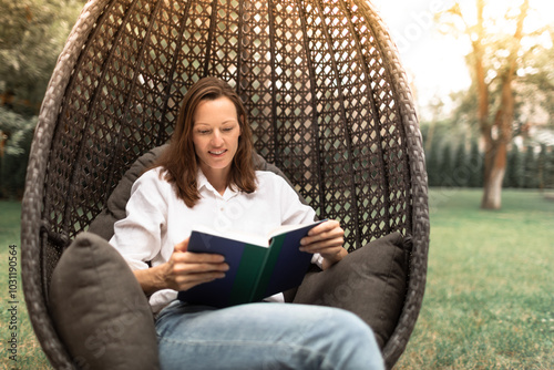 Young woman reading a book relaxing in a comfy lounge chair in a green nature setting enjoying weekend holiday 