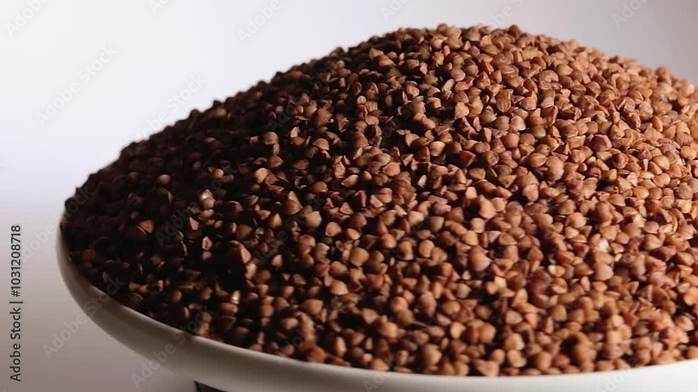 Dry Uncooked Brown Buckwheat Groats Heap on White Plate Rotating against White Background. Raw Large Buckwheat Grains. Russian, Ukrainian and Belarusian Culture Dry Uncooked Brown Buckwheat Groats Heap on White Plate Rotating against White Background. Raw Large Buckwheat Grains. Russian, Ukrainian and Belarusian Culture