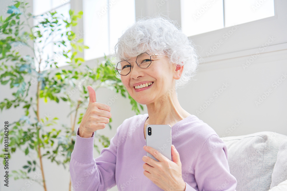 スマートフォンを持ってグッドサインするシニア女性 リビングルーム スマートフォンを持ってグッドサインするシニア女性 リビングルーム